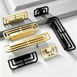 Könnte beinhalten: Eine Sammlung rechteckiger Schrankgriffe und Knöpfe in gebürstetem Gold und mattschwarzer Ausführung. Jedes Stück weist ein geometrisches Design mit offenem Rahmen und konzentrischen rechteckigen Linien auf. Einige tragen den Namen Evan Malik.