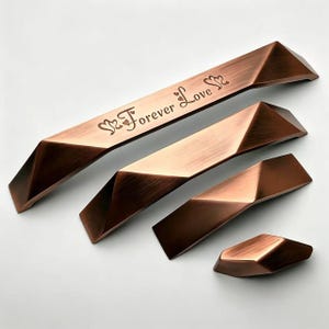 Pode incluir: Quatro puxadores geométricos para armários com acabamento em cobre escovado, dispostos por tamanho. O maior apresenta o texto gravado Forever Love com ícones de coração. Cada peça tem um design angular facetado com uma superfície metálica.