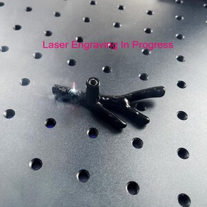 May include: A black, branch-shaped object with a vertical cylindrical attachment is undergoing laser engraving. The text "Laser Engraving In Progress" is visible above the object, which features a small, glowing laser point and smoke.