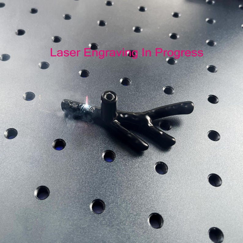 May include: A black, branch-shaped object with a vertical cylindrical attachment is undergoing laser engraving. The text "Laser Engraving In Progress" is visible above the object, which features a small, glowing laser point and smoke.
