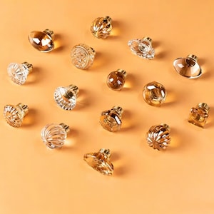 May include: A collection of fifteen decorative cabinet knobs featuring various shapes and textures. The knobs are crafted from clear and amber-coloured glass or acrylic, each mounted on a small metallic base. The designs include faceted, ribbed, and smooth circular or geometric forms.