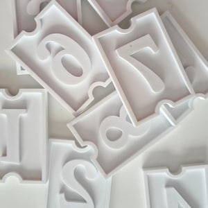 May include: White, die-cut number tiles, featuring the numbers 1, 2, 4, 6, 7, and 8. The numbers are in a sans-serif font and are raised from the tile surface. The tiles have a decorative border and a notch at the top.