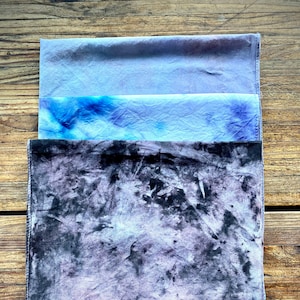 May include: Three square fabric squares, one black with white and gray marbling, one light blue with darker blue marbling, and one solid light blue. The black square has a blue tag with the text "WOVEN DREAMS" on it.