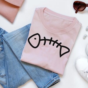 May include: A pink t-shirt with a black fish bone graphic. The shirt is laid out on a white surface with blue jeans and a pink purse.