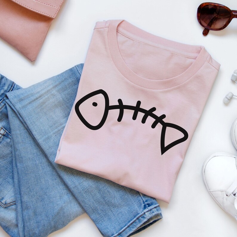 May include: A pink t-shirt with a black fish bone graphic. The shirt is laid out on a white surface with blue jeans and a pink purse.