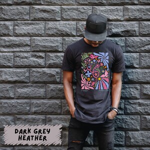 K&ouml;nnte beinhalten: Ein dunkelgraues T-Shirt mit einem bunten Grafikdesign mit medizinischen Symbolen und Blumen. Das Design enth&auml;lt ein Stethoskop, Verb&auml;nde, Pillen, eine Spritze und Blumen. Der Text "Compassion" und "Medicine 2021" ist ebenfalls im Design enthalten.