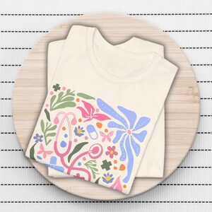 K&ouml;nnte beinhalten: Ein wei&szlig;es T-Shirt mit einem farbenfrohen Blumendesign, das ein Stethoskop, Pillen und andere medizinische Symbole zeigt. Das Design ist in den Farben Rosa, Blau, Gr&uuml;n und Orange gehalten.