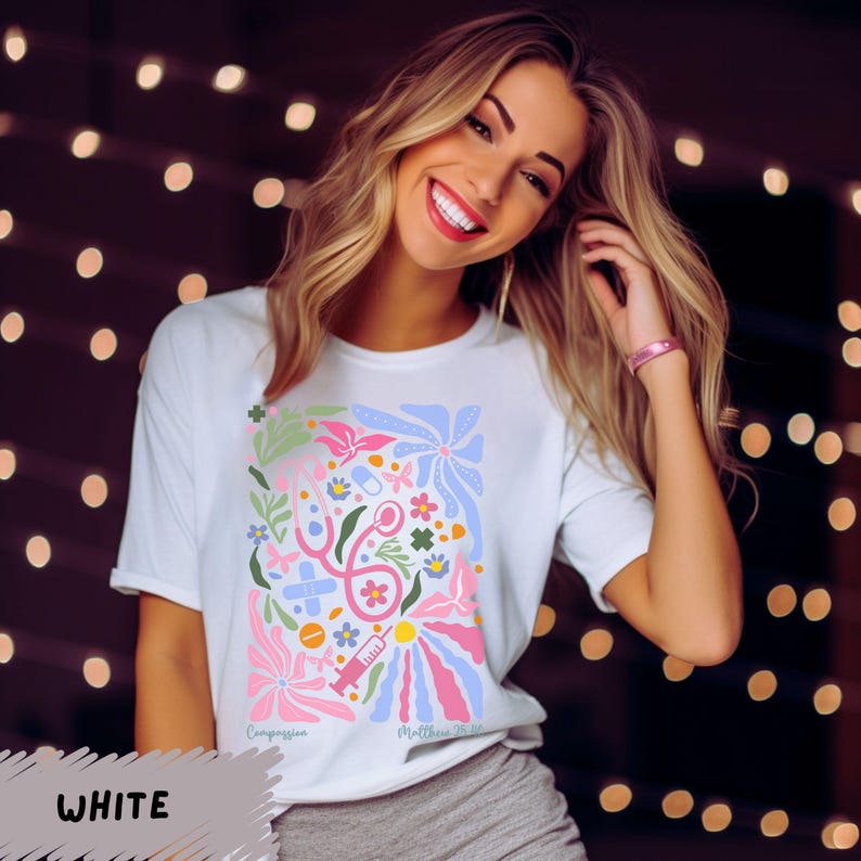 K&ouml;nnte beinhalten: Wei&szlig;es T-Shirt mit einem farbenfrohen Grafikdesign mit medizinischen Symbolen und Blumen. Der Text "Compassion" und "Medicine That Heals" ist auf dem Shirt aufgedruckt.