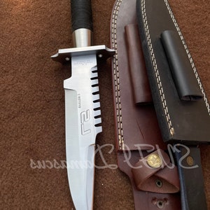 Può includere: Un coltello argentato con manico nero e fodero in pelle. Il coltello ha un bordo seghettato e il testo "L.T.2" è stampato sulla lama. Il fodero ha una cinghia in pelle marrone con una fibbia in ottone.