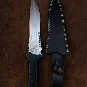 May include: A black-handled knife with a silver blade and a serrated edge. The blade has a circular emblem with the text "S.R.A.T.E.  KOO33AЯ  POLICE DES" on it. The knife is in a black leather sheath with a stitched edge.