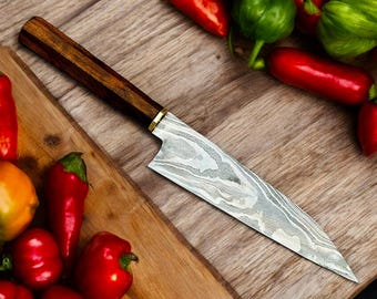 May include: A close-up of a kitchen knife with a wooden handle and a patterned blade. The knife is lying on a wooden cutting board with red and green peppers.