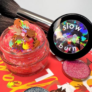 Puede incluir: Un tarro de vidrio transparente lleno de purpurina roja y confeti con forma de llama, junto a un recipiente de maquillaje negro con la inscripción "slow burn". Dos brochas de maquillaje están en el fondo. Se ven otros productos de maquillaje.