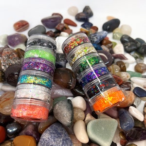 May include: Two clear plastic containers filled with colorful glitter. Each container has six layers of glitter in different colors, including green, purple, gold, and orange. The containers are sitting on a bed of colorful rocks.
