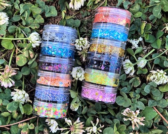 Op de afbeelding: Twee doorzichtige plastic potten gevuld met lagen kleurrijke glitter. Elke pot heeft zes lagen glitter in verschillende kleuren, waaronder roze, blauw, goud en zwart.