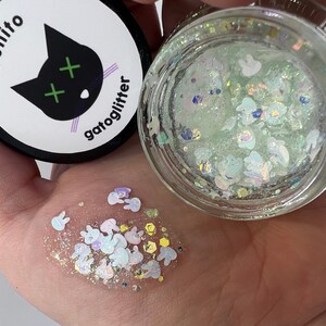 Może przedstawiać: Mały słoiczek zielonego brokatu z białymi kształtami kr&oacute;lik&oacute;w i innymi tęczowymi brokatami. Słoiczek jest oznaczony "benito gatoglitter" z czarnym kotem z zielonymi X jako oczami.