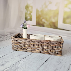 May include: A woven wicker basket with two rolls of white toilet paper and a white vase with purple and pink flowers.