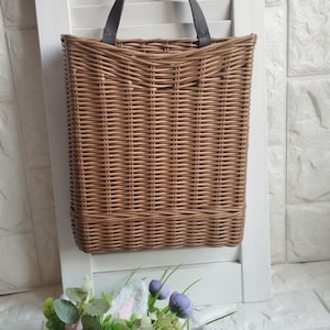 May include: A rectangular woven basket in shades of brown, with a black handle, hangs from a metal hook. The basket is mounted on a white shutter. A floral arrangement with purple and pink flowers and green leaves is in the foreground.
