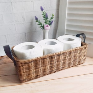 May include: A woven wicker basket with black leather handles holds three rolls of white toilet paper. A small vase with a single pink flower and purple sprigs sits behind the basket.