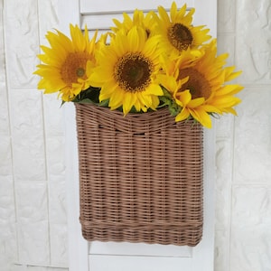 May include: A brown wicker basket filled with bright yellow sunflowers, mounted on a white wooden shutter. The sunflowers have large, dark brown centers and vibrant yellow petals. The basket is rectangular and has a woven texture.