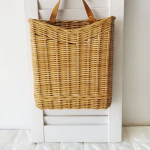 Op de afbeelding: Rechthoekige rieten mand met een leren handvat, hangend aan een witte luik. De mand is gemaakt van lichtbruin vlechtwerk en is ontworpen voor wandmontage. De luik is wit met horizontale lamellen.