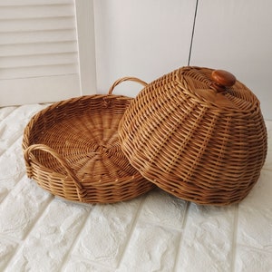 Könnte beinhalten: Ein rundes Serviertablett aus Weide mit einem Kuppeldeckel. Das Tablett hat zwei Griffe und der Deckel hat einen Holzknopf.