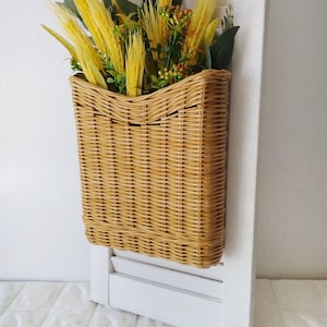 May include: A woven wicker basket filled with yellow and green decorative plants, mounted on a white shutter. The basket is rectangular and has a natural, light brown color. The plants add a touch of color.