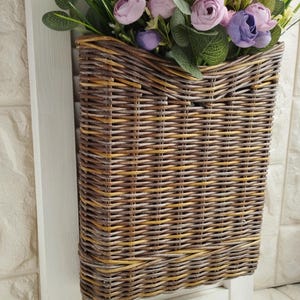 May include: A rectangular woven wicker basket with a white wooden frame, filled with faux flowers in shades of pink and purple, and green foliage. The basket is designed for wall mounting.