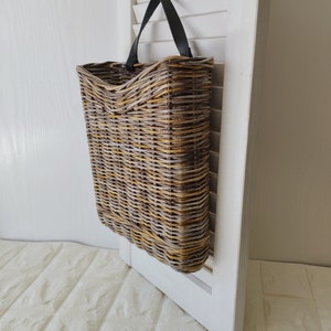 Op de afbeelding: Geweven rieten mand met een rechthoekige vorm en een zwart leren handvat, hangend aan een witte luik. De mand heeft een natuurlijke kleur met tinten bruin en geel.