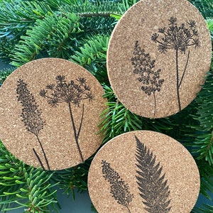 Könnte beinhalten: Drei runde Untersetzer aus Kork mit schwarzen botanischen Designs, darunter Farne und andere Pflanzen. Die Untersetzer sind auf einem Bett aus grünen Tannennadeln angeordnet, was eine natürliche, erdige Ästhetik erzeugt.