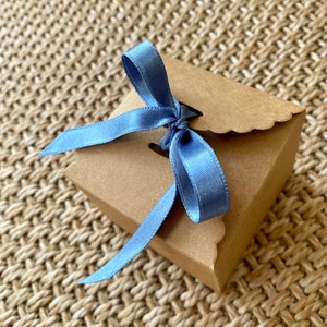 Könnte beinhalten: Eine kleine, quadratische, braune Geschenkbox mit gewelltem Rand, gebunden mit einem hellblauen Satinband. Die Box ist mit einer Schleife verschlossen. Die Box liegt auf einer gewebten, beigefarbenen Oberfläche.