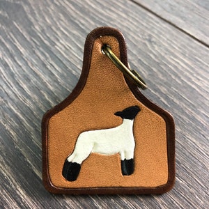 May include: A brown leather keychain shaped like a livestock tag with a white lamb silhouette on a tan background.