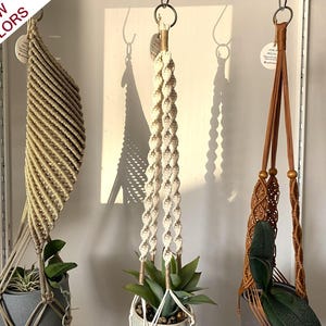 Puede incluir: Tres colgadores de plantas de macramé suspendidos de anillos de metal. El colgador izquierdo es de tono dorado con patrones de cuerda diagonales, el central es de color crema con un diseño de nudos trenzados y el derecho es marrón.