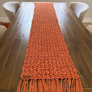 May include: A long, rectangular table runner in a vibrant burnt orange colour, featuring a thick, hand-knotted macrame texture. The runner is finished with long, straight fringe tassels at both ends and includes a small, rectangular brown leather tag.