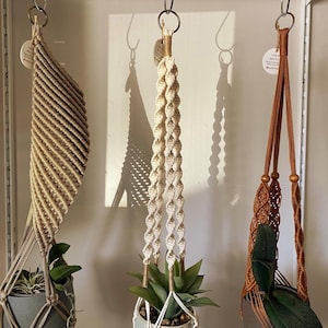 Op de afbeelding: Drie macramé plantenhangers in verschillende stijlen. De eerste is een bruin, ruitvormig ontwerp. De tweede is een crèmekleurige, gedraaide touwstijl. De derde is een bruin, geweven ontwerp met houten kralen. Elke hanger bevat een potplant.