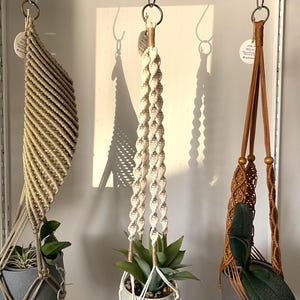 Puede incluir: Tres colgadores de plantas de macramé suspendidos de anillos de metal. El colgador izquierdo es de color tostado con tejido diagonal, el central es crema con diseño de cuerda trenzada y el derecho es marrón con patrón de rombos.