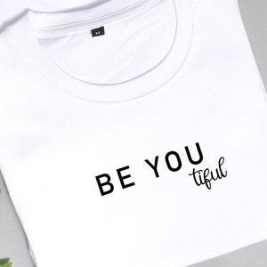 Könnte beinhalten: Weißes T-Shirt mit dem Aufdruck "BE YOU tiful" in Schwarz.