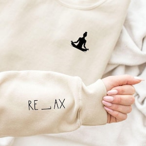Könnte beinhalten: Ein beiger Pullover mit einer schwarzen Silhouette einer Person in einer Yoga-Pose auf der Vorderseite und dem Wort "RE_LAX" auf dem Ärmel.