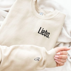 Könnte beinhalten: Ein beiger Pullover mit dem Wort "Liebs." auf der Vorderseite und einem kleinen Herz auf dem Ärmel gestickt.