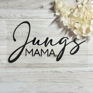 Könnte beinhalten: Schwarzes, lasergeschnittenes Schild mit den Worten "Jungs MAMA" in einer Schreibschrift. Das Schild befindet sich auf einem weiß-grauen Holzmaserungshintergrund, mit weißen Blumen in der oberen rechten Ecke. Ein dekorativer Artikel für ein Zuhause.