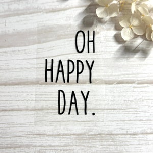 Könnte beinhalten: Ein transparenter Aufkleber mit schwarzem Text, der "OH HAPPY DAY." lautet.