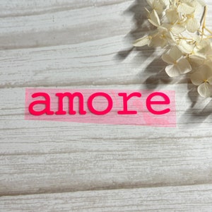 Könnte beinhalten: Rosa Vinylbuchstaben, die das Wort "amore" auf einem hellen Holzhintergrund buchstabieren.