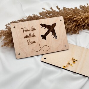 K&ouml;nnte beinhalten: Ein Holzschild mit dem Text "F&uuml;r die n&auml;chste Reise" und einem ausgeschnittenen Flugzeug. Das Schild ist hellbraun und hat zwei goldene Stecknadeln.