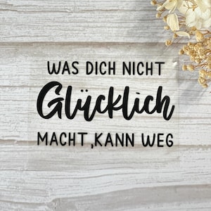 Könnte beinhalten: Ein klarer Aufkleber mit schwarzem Text, der "WAS DICH NICHT Glücklich MACHT, KANN WEG" auf einem weißen Holzgrund liest. Getrocknete Blumen befinden sich in der oberen rechten Ecke.