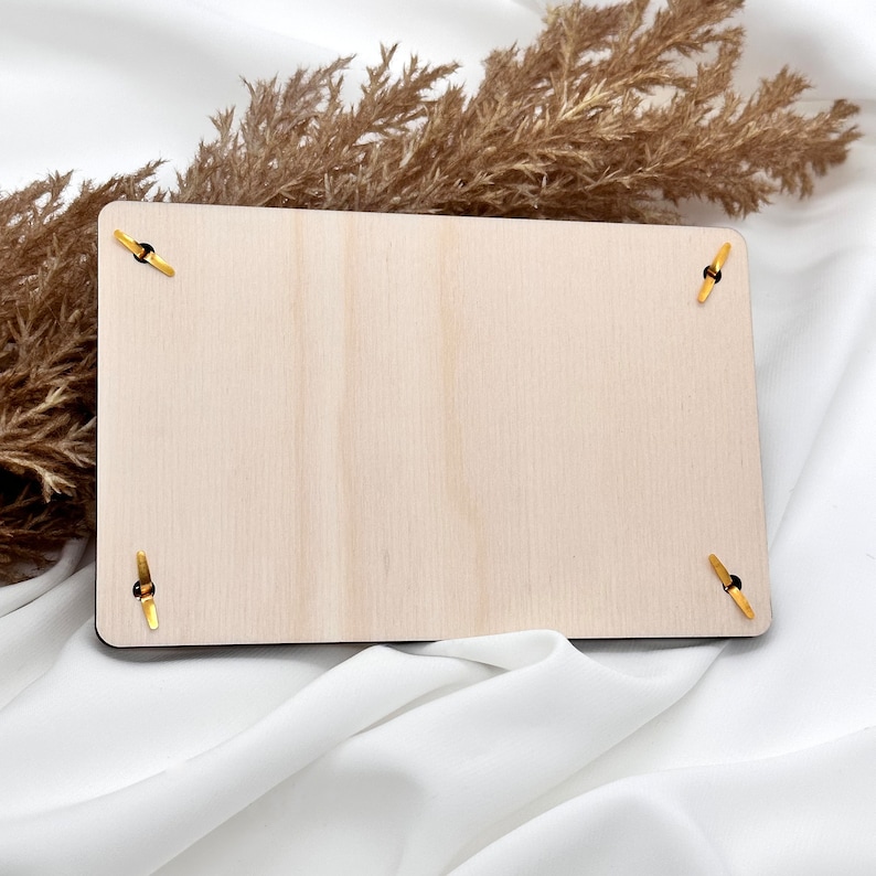 K&ouml;nnte beinhalten: Ein leeres Holzschild mit vier goldenen Metallecken. Das Schild liegt auf einer wei&szlig;en Stoffoberfl&auml;che.