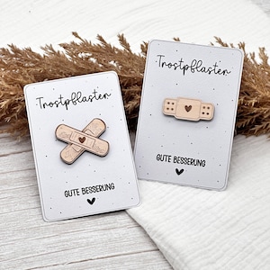 Könnte beinhalten: Zwei Holz-Pflaster-Anstecknadeln mit Herzdetails auf weißen Karten. Die Karten tragen die Aufschriften "Trostpflaster" und "Gute Besserung" mit einem Herzsymbol. Die Anstecknadeln sind hellbraun.