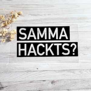Könnte beinhalten: Zwei schwarze rechteckige Schilder mit weißem Text, der "SAMMA" und "HACKTS?" lautet, werden auf einer hellen Holzmaserungsoberfläche angezeigt. Die Schilder bestehen aus einem transparenten Material. Getrocknete Blumen befinden sich in der oberen linken Ecke.