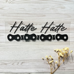 Könnte beinhalten: Schwarzer Vinyl-Aufkleber mit dem Wort "Hätte" zweimal, über einem Ketten-Design. Der Text ist in einer Schreibschrift. Der Aufkleber befindet sich auf einer transparenten Rückseite, mit einem Holz-Hintergrund.