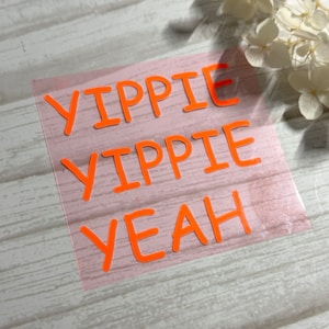 Könnte beinhalten: Ein rosa Aufkleber mit den Worten "YIPPIE YIPPIE YEAH" in orange.