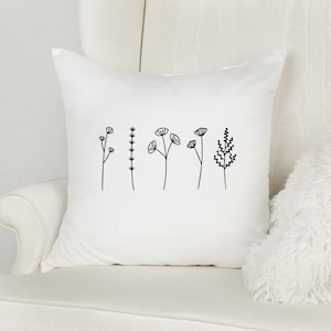 Könnte beinhalten: Quadratisches weißes Kissen mit einem minimalistischen Blumendesign in Schwarz. Das Design zeigt verschiedene stilisierte Blumen und Pflanzen. Das Kissen liegt auf einem weißen Stuhl, mit einem flauschigen weißen Kissen im Hintergrund.