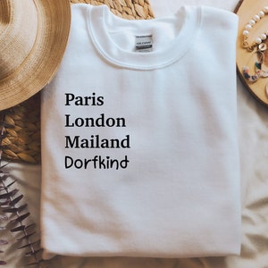 Könnte beinhalten: Weißer Pullover mit schwarzem Text, der "Paris London Mailand Dorfkind" lautet.